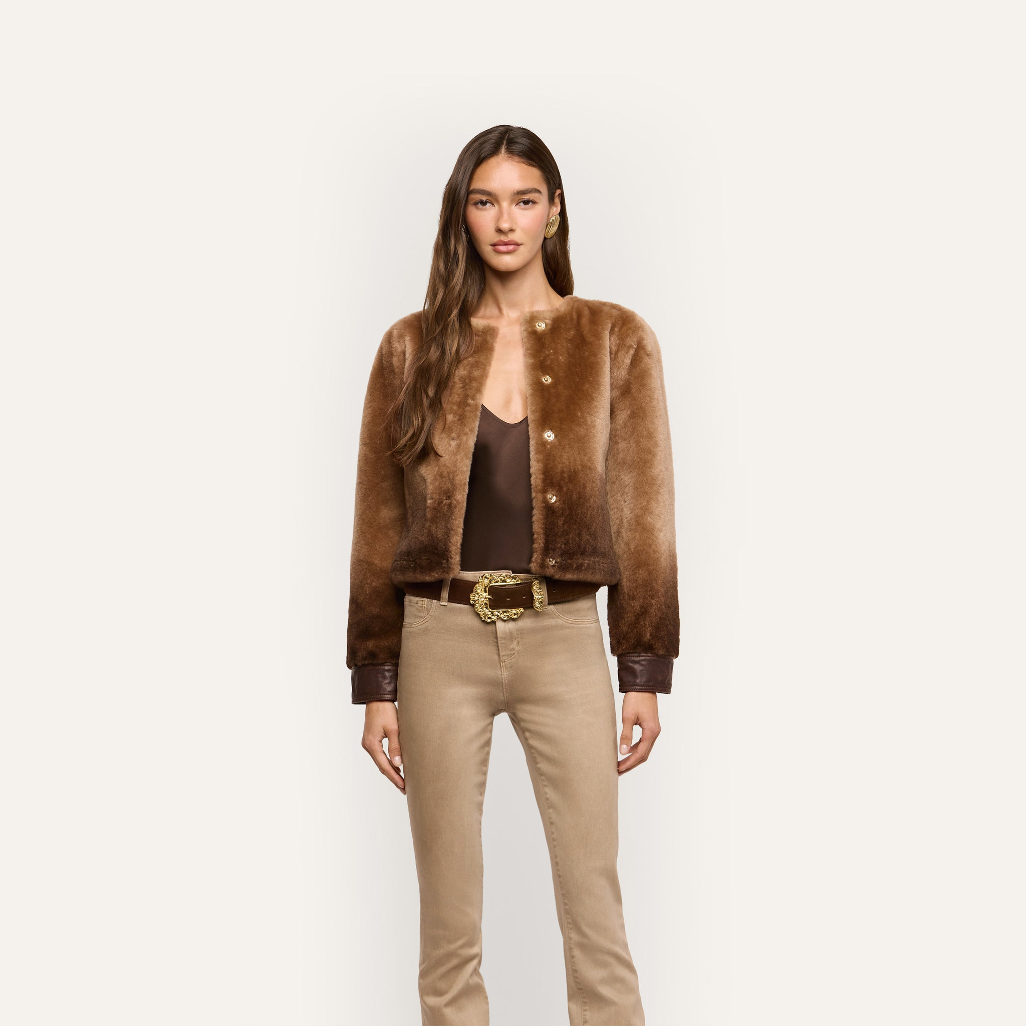 A model wears a brown ombré Voltaire Shearling Jacket over a dark brown camisole and tan trousers with a gold-buckled belt.