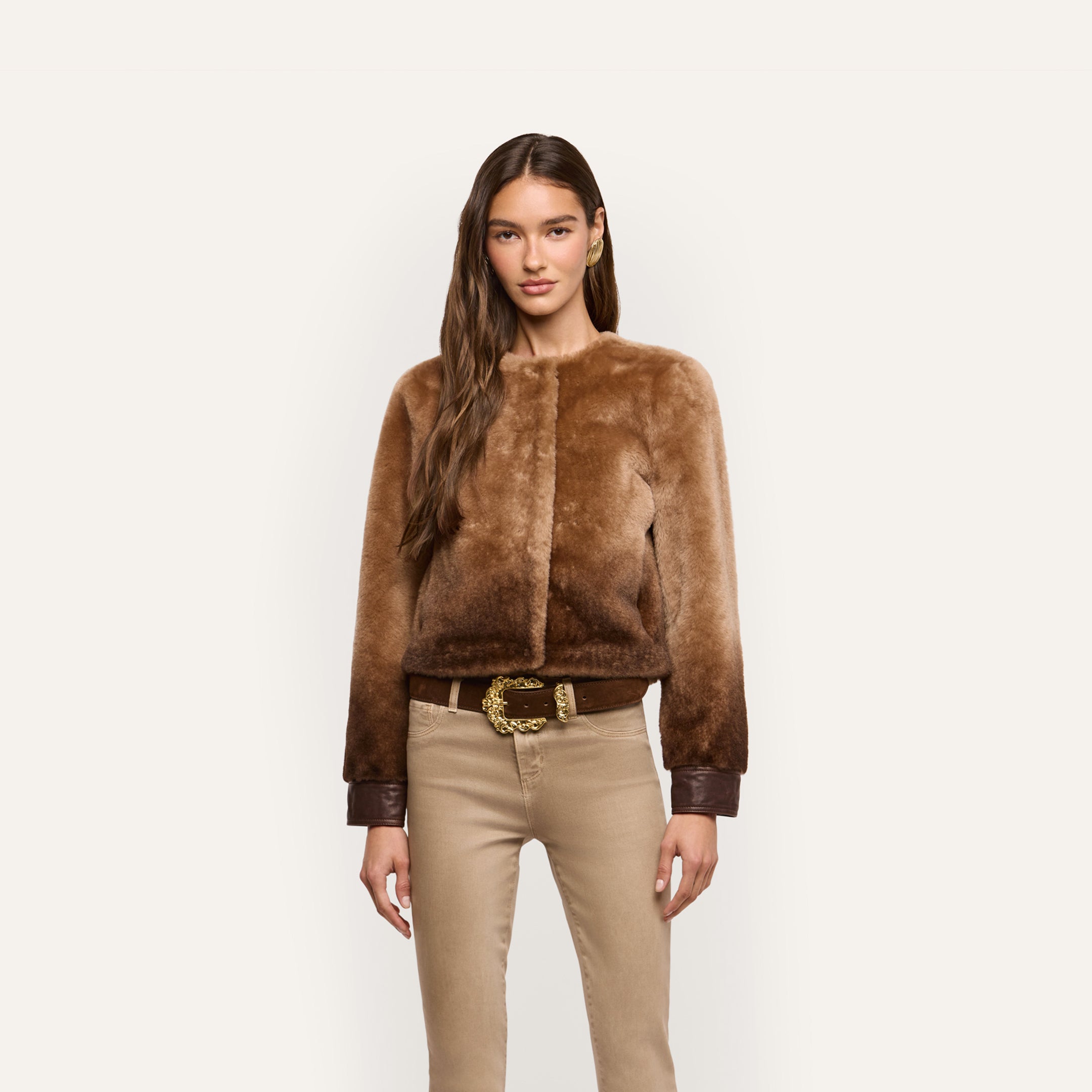 A woman wearing a brown ombré merino shearling Voltaire Shearling Jacket with leather cuffs over tan pants and a belt with a gold-toned buckle.
