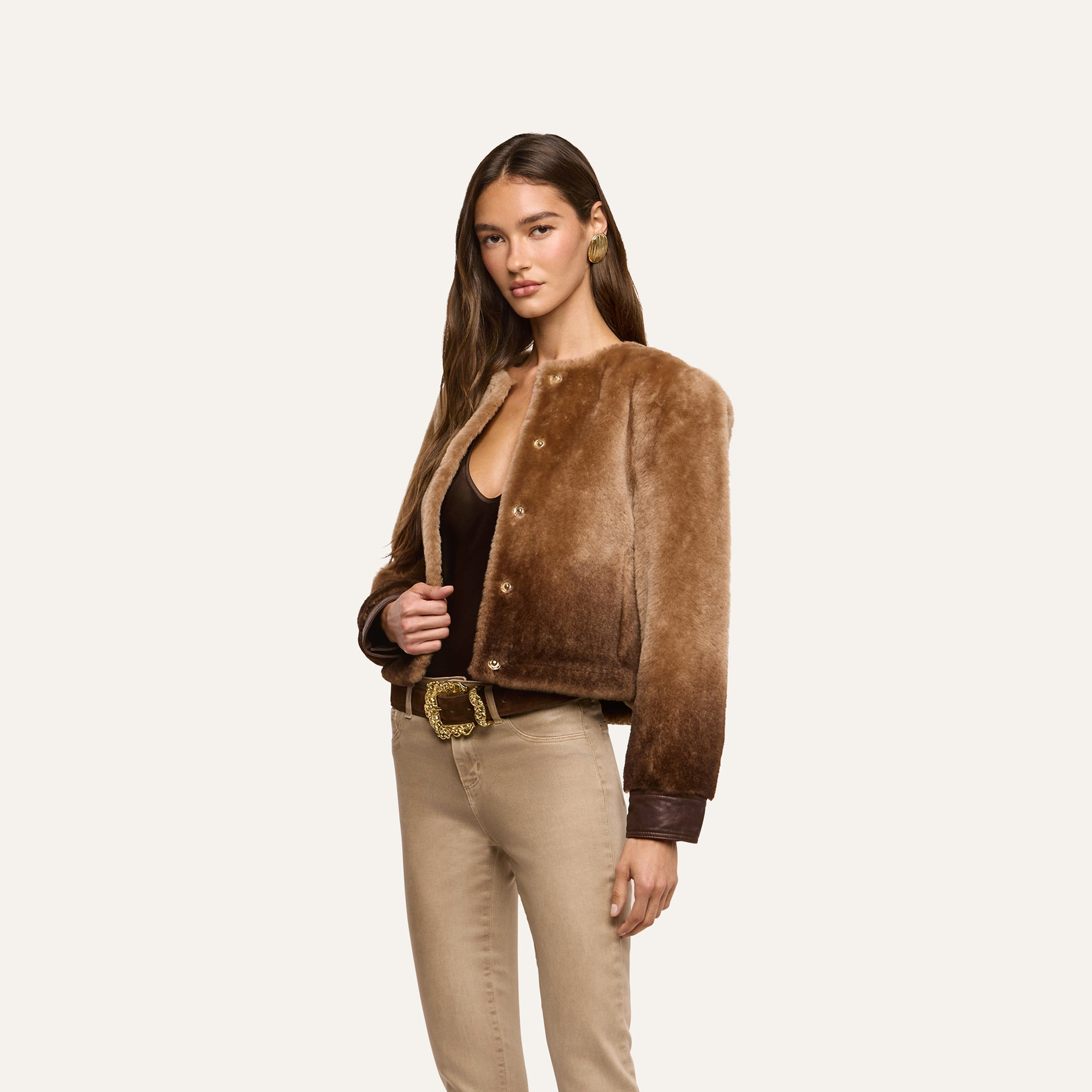 A woman wearing a brown ombré Voltaire Shearling Jacket, a dark bodysuit, and tan jeans with a gold-buckle belt.