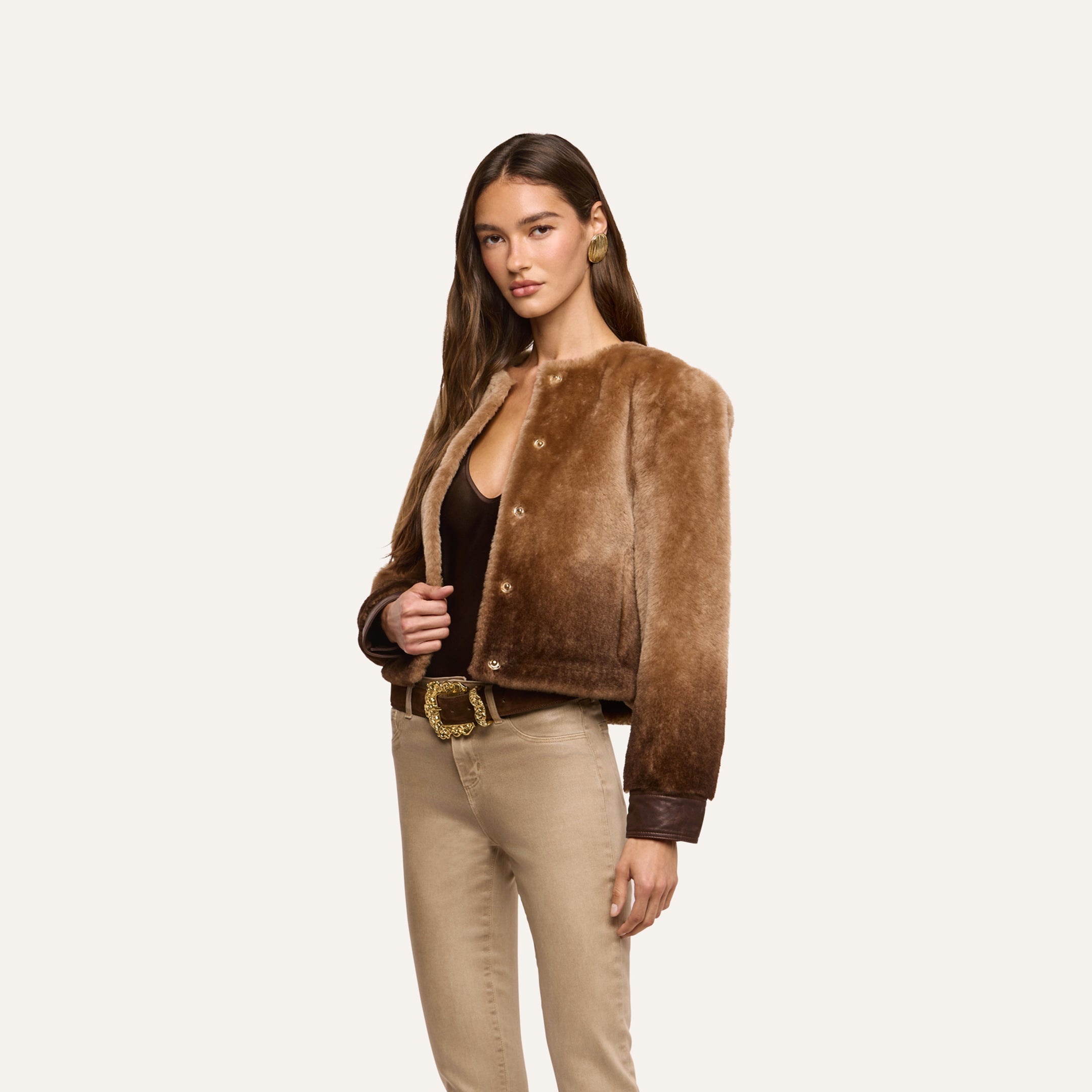 A woman wearing a brown ombré Voltaire Shearling Jacket, a dark bodysuit, and tan jeans with a gold-buckle belt.