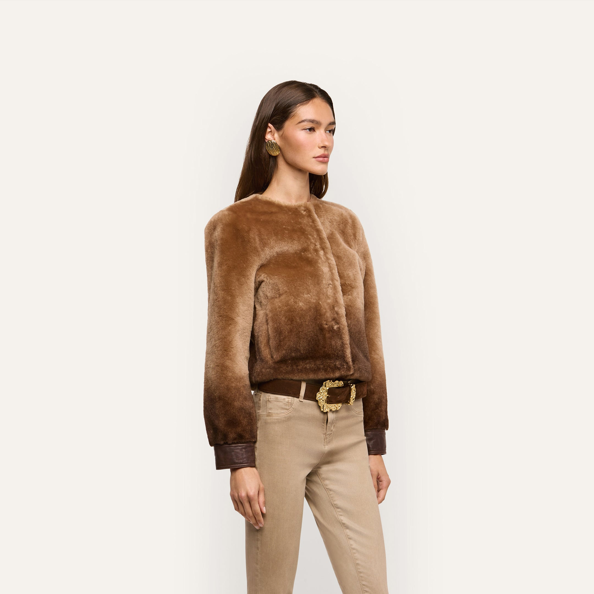 A woman wears a brown cropped Voltaire Shearling Jacket with dark brown leather cuffs over tan jeans and a brown belt with an ornate gold buckle.
