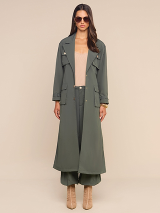 Wearing the Axel Windbreaker Trench styled as a sleek olive green utility jacket, paired with matching wide-leg pants, a beige top, tan heeled boots, and sunglasses, the woman stands facing forward against a plain beige background.
