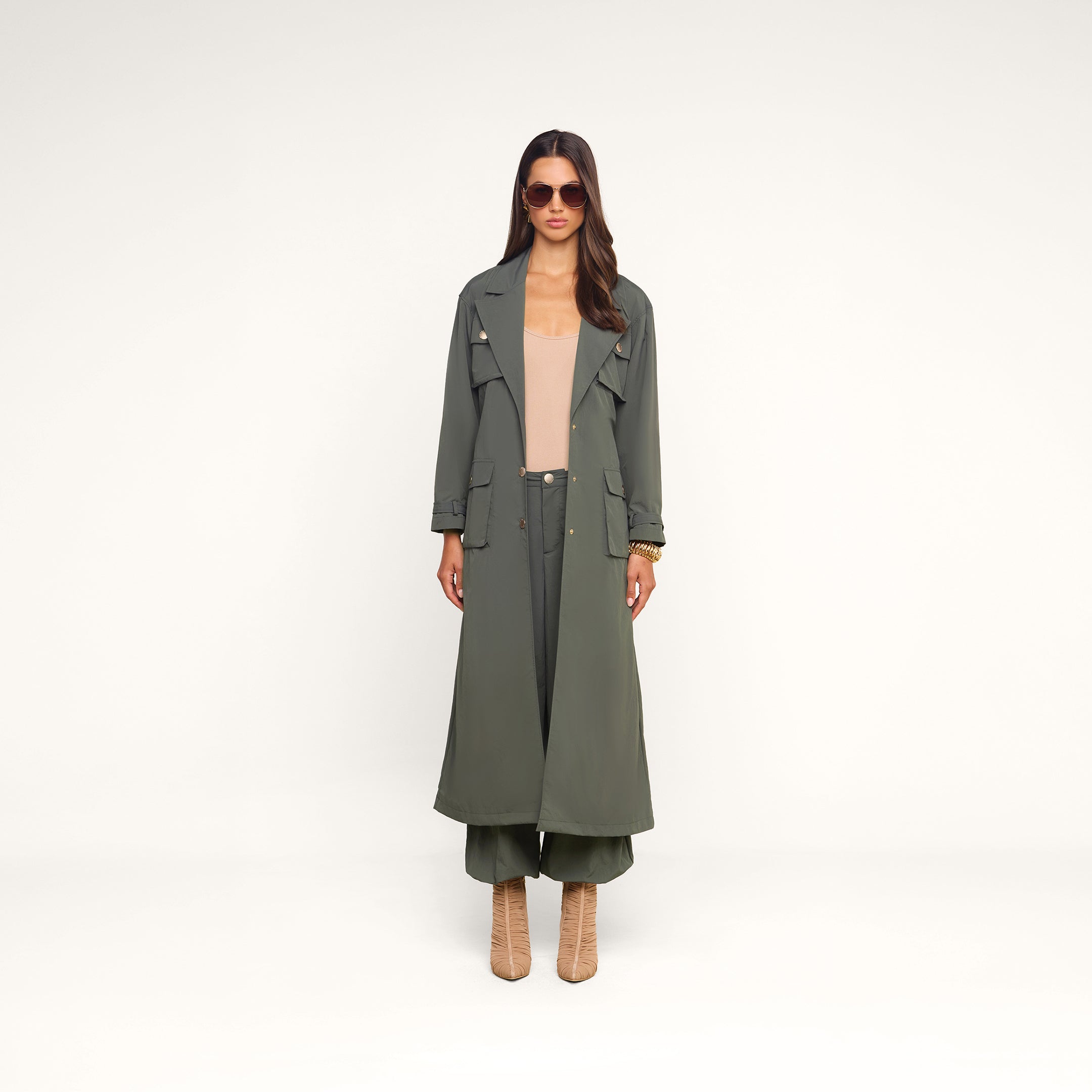 Wearing the Axel Windbreaker Trench styled as a sleek olive green utility jacket, paired with matching wide-leg pants, a beige top, tan heeled boots, and sunglasses, the woman stands facing forward against a plain beige background.