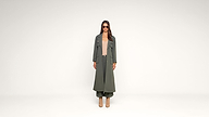 A woman stands against a plain white background wearing a long olive green trench coat, matching pants, beige top, heeled boots, and sunglasses.