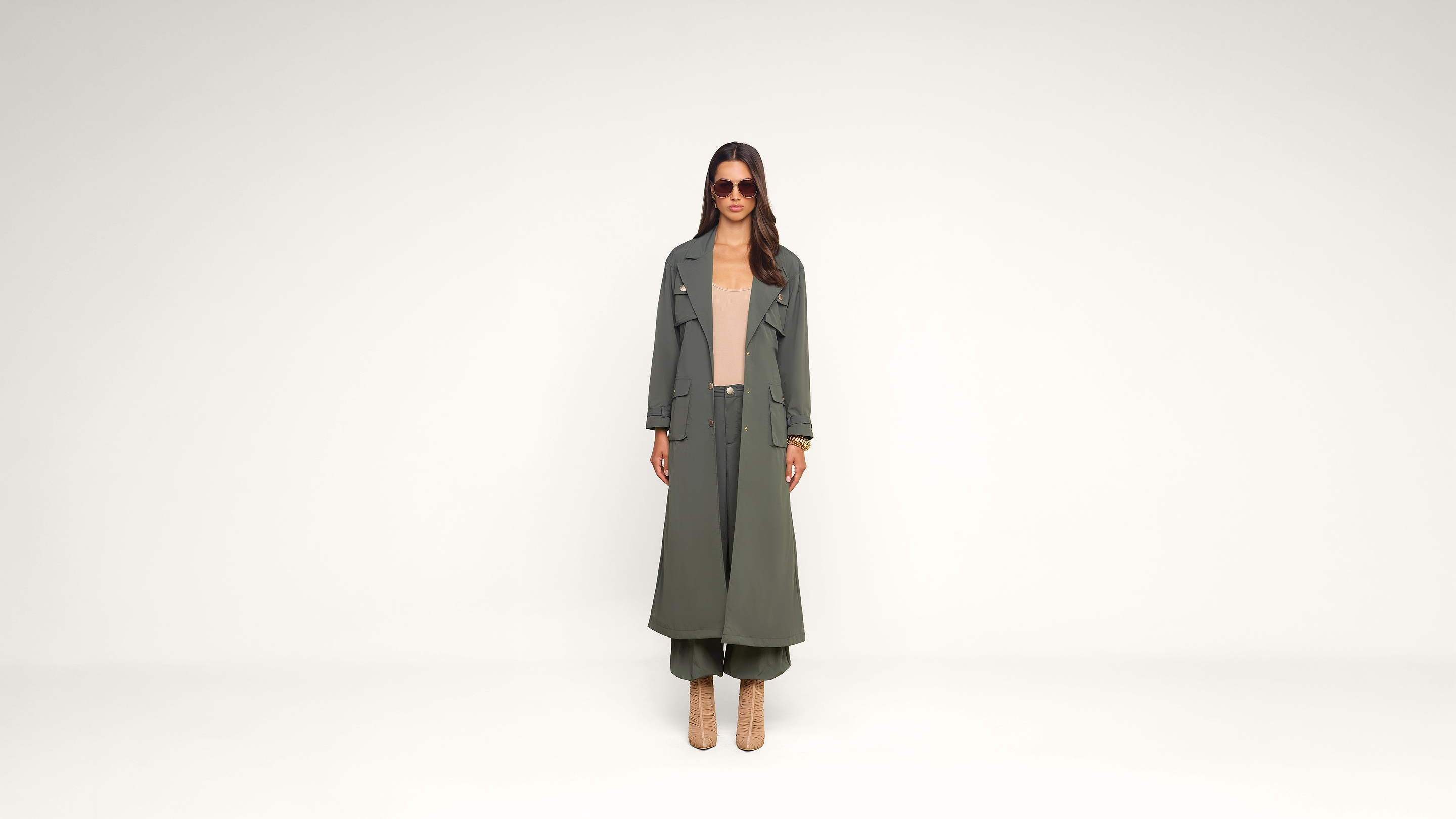 A woman stands against a plain white background wearing a long olive green trench coat, matching pants, beige top, heeled boots, and sunglasses.