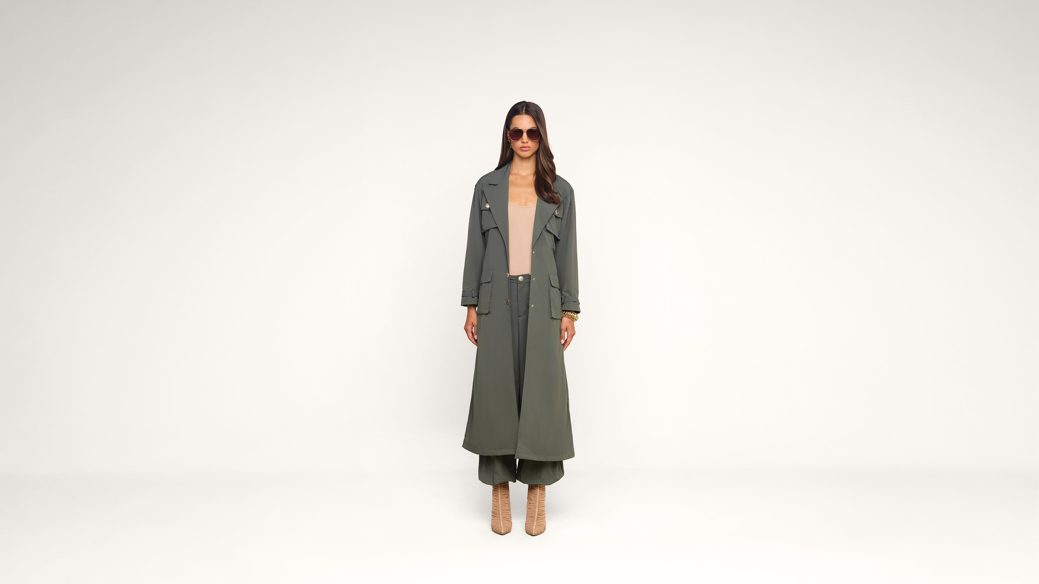 A woman stands against a plain white background wearing a long olive green trench coat, matching pants, beige top, heeled boots, and sunglasses.