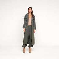 A woman with long dark hair and a neutral expression stands against a beige background wearing the Axel Windbreaker Trench in olive green, styled with wide-leg pants, a beige top, and tan heeled boots.
