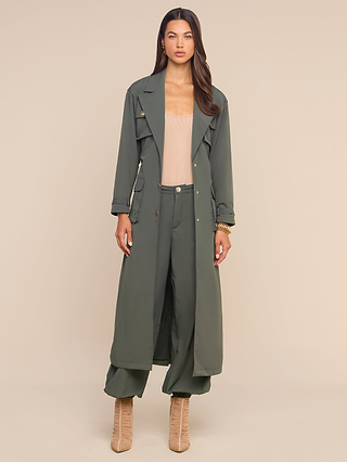 A woman with long dark hair and a neutral expression stands against a beige background wearing the Axel Windbreaker Trench in olive green, styled with wide-leg pants, a beige top, and tan heeled boots.
