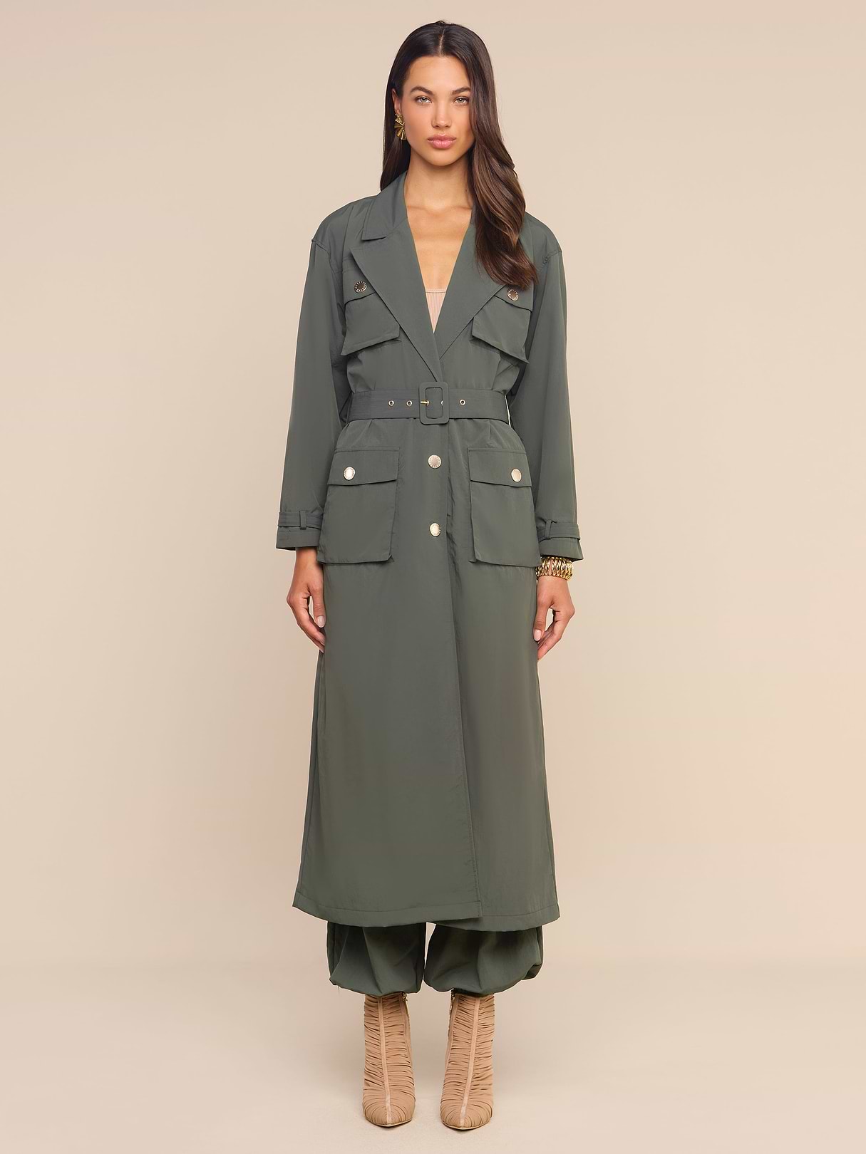 A woman stands facing forward, wearing the Axel Windbreaker Trench in olive green over matching pants and tan heeled boots, against a plain white background.
