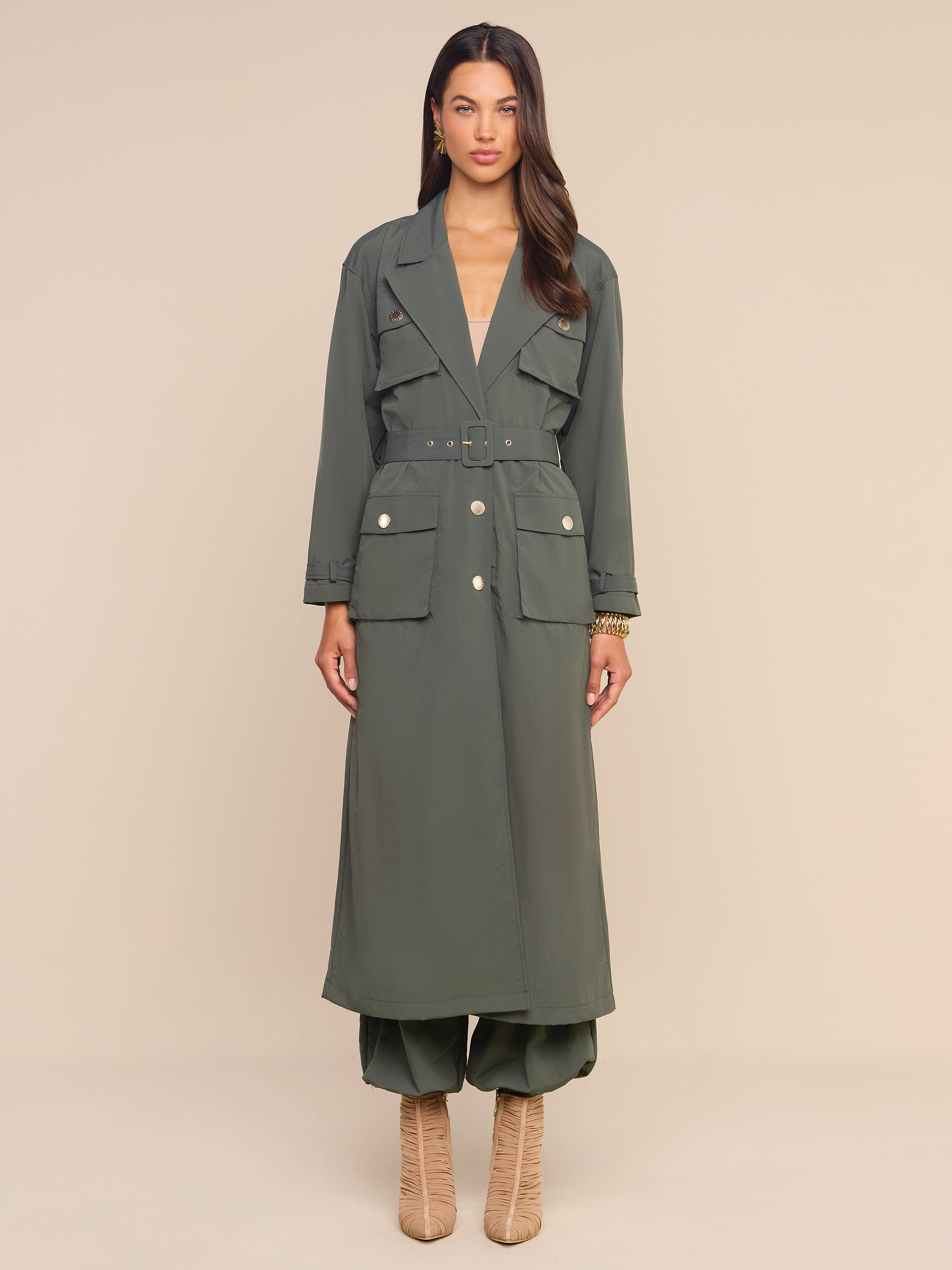 A woman stands against a plain beige background wearing the Axel Windbreaker Trench—an olive green, long belted trench coat with large front pockets reminiscent of a stylish utility jacket—paired with matching wide-leg pants and beige heeled boots.
