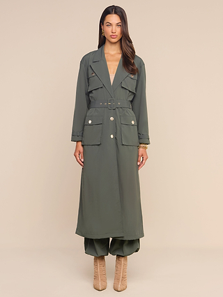 A woman stands against a plain beige background wearing the Axel Windbreaker Trench—an olive green, long belted trench coat with large front pockets reminiscent of a stylish utility jacket—paired with matching wide-leg pants and beige heeled boots.