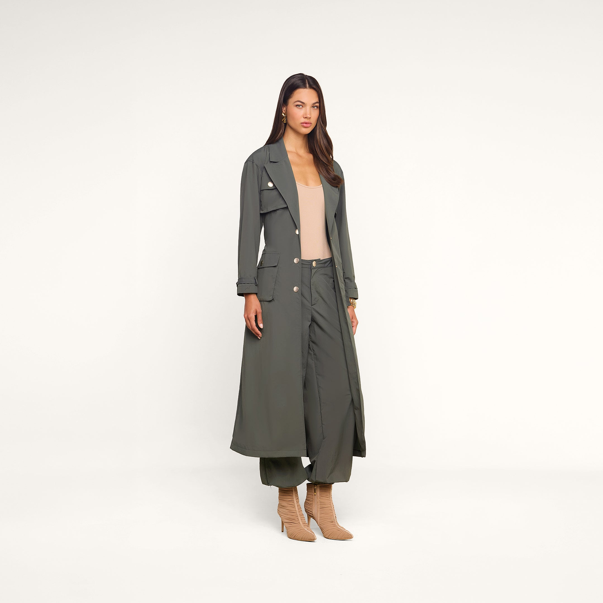 Wearing the Axel Windbreaker Trench, a woman stands against a neutral background, paired with matching wide-leg pants, a beige top, and tan heeled boots.