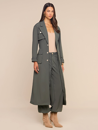 Wearing the Axel Windbreaker Trench, a woman stands against a neutral background, paired with matching wide-leg pants, a beige top, and tan heeled boots.