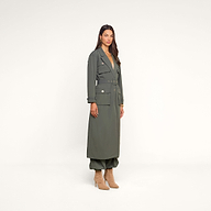 A woman stands against a light beige background wearing the Axel Windbreaker Trench, a long belted olive green coat with large utility-style pockets, paired with matching pants and beige heeled boots.