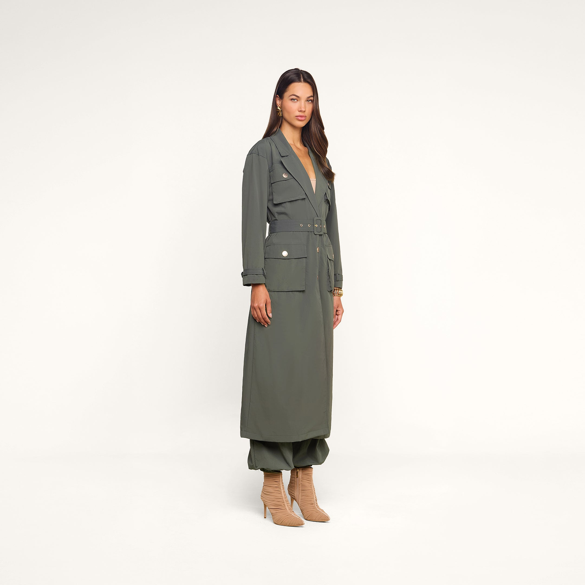 A woman stands against a light beige background wearing the Axel Windbreaker Trench, a long belted olive green coat with large utility-style pockets, paired with matching pants and beige heeled boots.