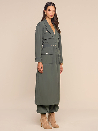 A woman stands against a light beige background wearing the Axel Windbreaker Trench, a long belted olive green coat with large utility-style pockets, paired with matching pants and beige heeled boots.