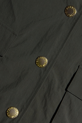 Close-up of the Axel Windbreaker Trench, a dark fabric utility jacket featuring large, round gold buttons engraved with “Burberry London England” and two rectangular front pockets.