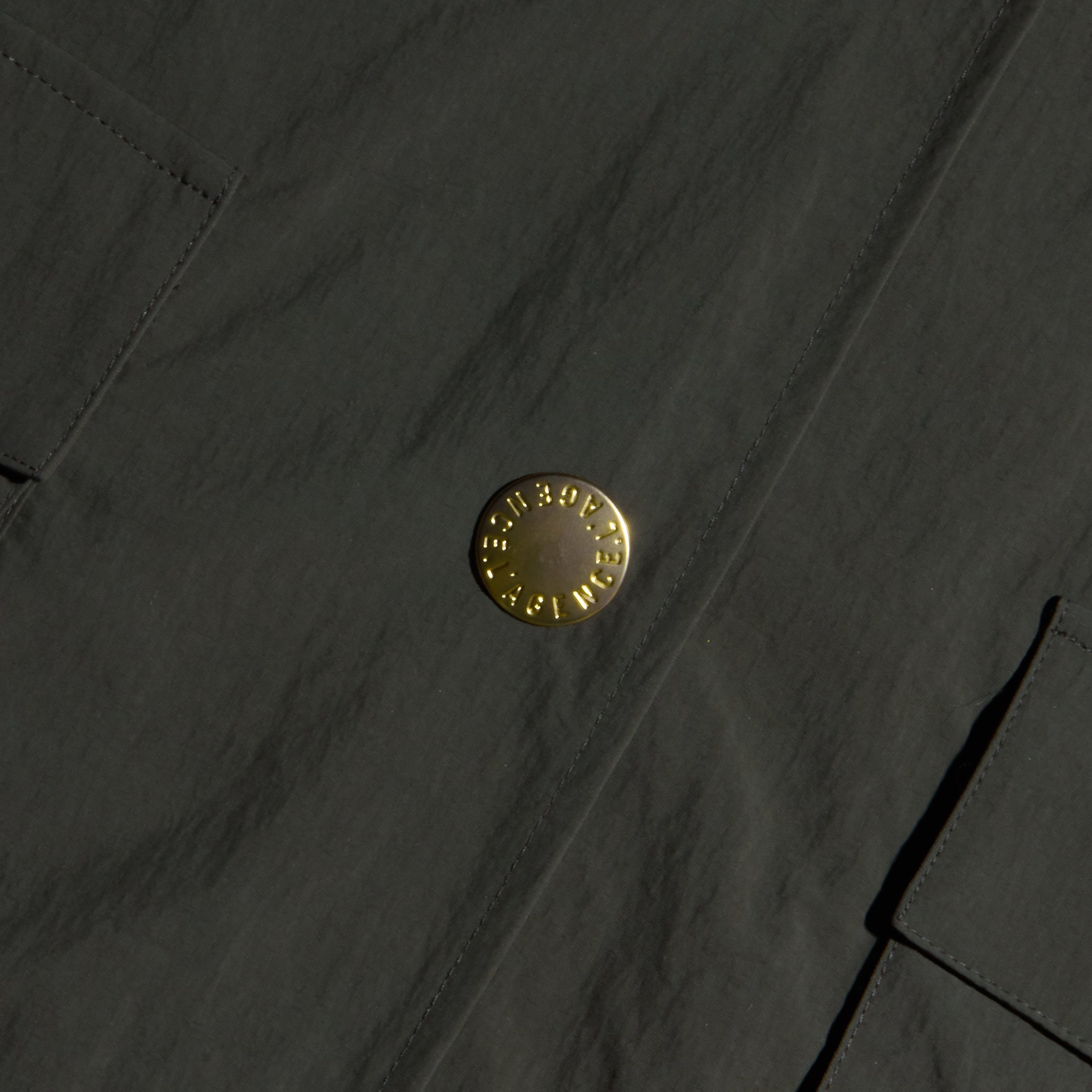 Close-up of the Axel Windbreaker Trench, a dark fabric utility jacket featuring large, round gold buttons engraved with “Burberry London England” and two rectangular front pockets.
