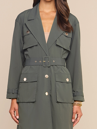 A person wearing the Axel Windbreaker Trench—a dark green belted trench coat featuring large buttons and multiple front pockets, inspired by classic utility jackets—stands against a plain beige background.