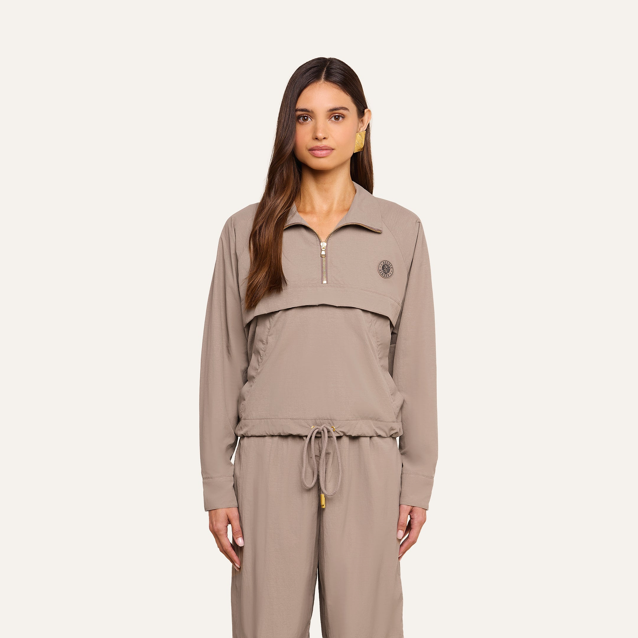 A woman with long brown hair wears the Vex Cropped Windbreaker in taupe, paired with matching drawstring pants, standing against a plain light background and looking directly at the camera with a neutral expression.