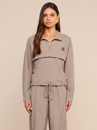 A woman with long brown hair wears the Vex Cropped Windbreaker, a taupe activewear jacket with a zippered collar, paired with matching drawstring pants as she stands against a beige background.