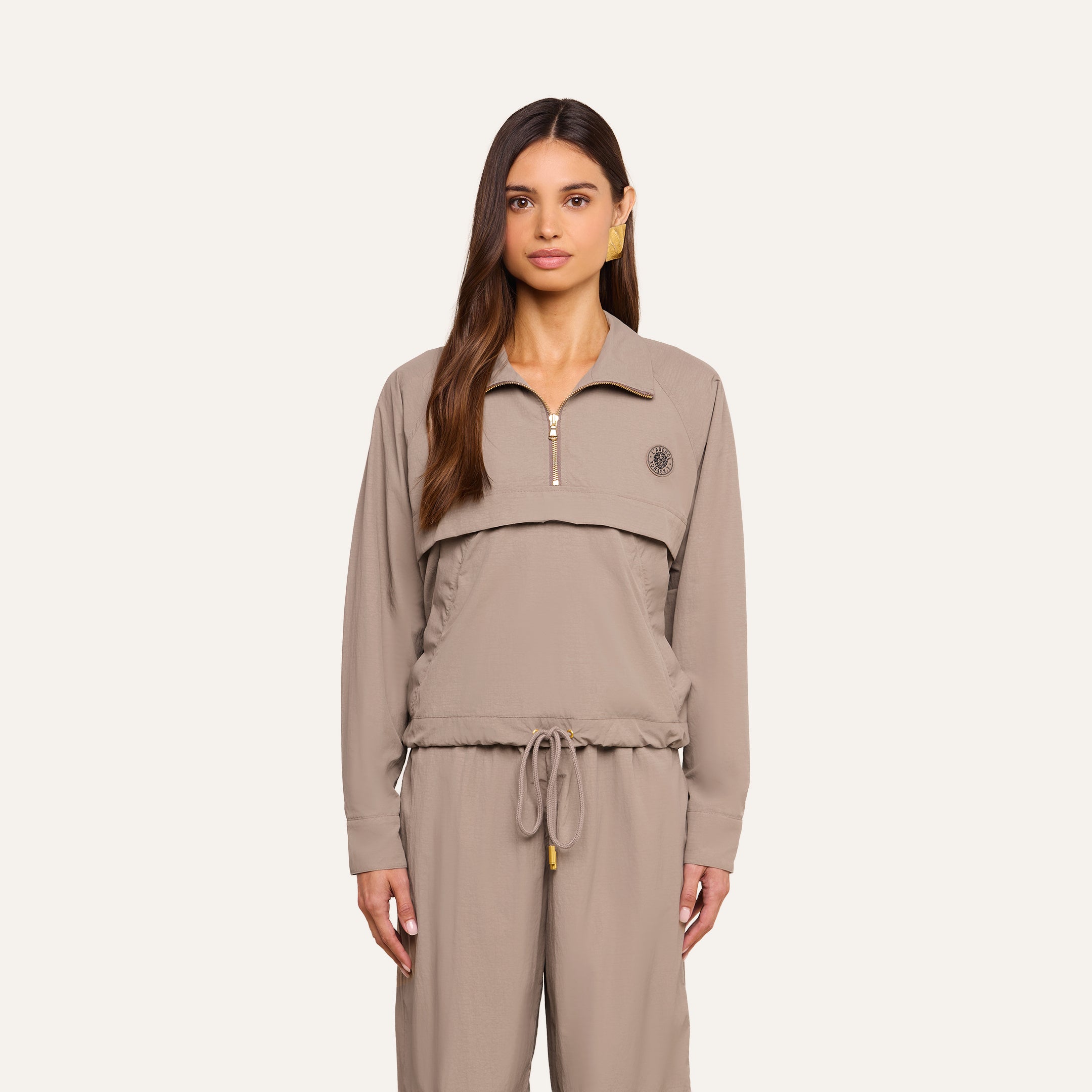 A woman with long brown hair wears the Vex Cropped Windbreaker in taupe, paired with matching drawstring pants, standing against a plain light background and looking directly at the camera with a neutral expression.