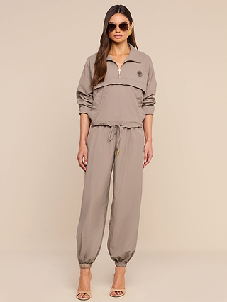 A woman stands against a beige background wearing the Vex Cropped Windbreaker and matching taupe tracksuit, featuring a zip-up top, drawstring waist, and jogger pants. She pairs the look with long hair, sunglasses, and beige heeled sandals.