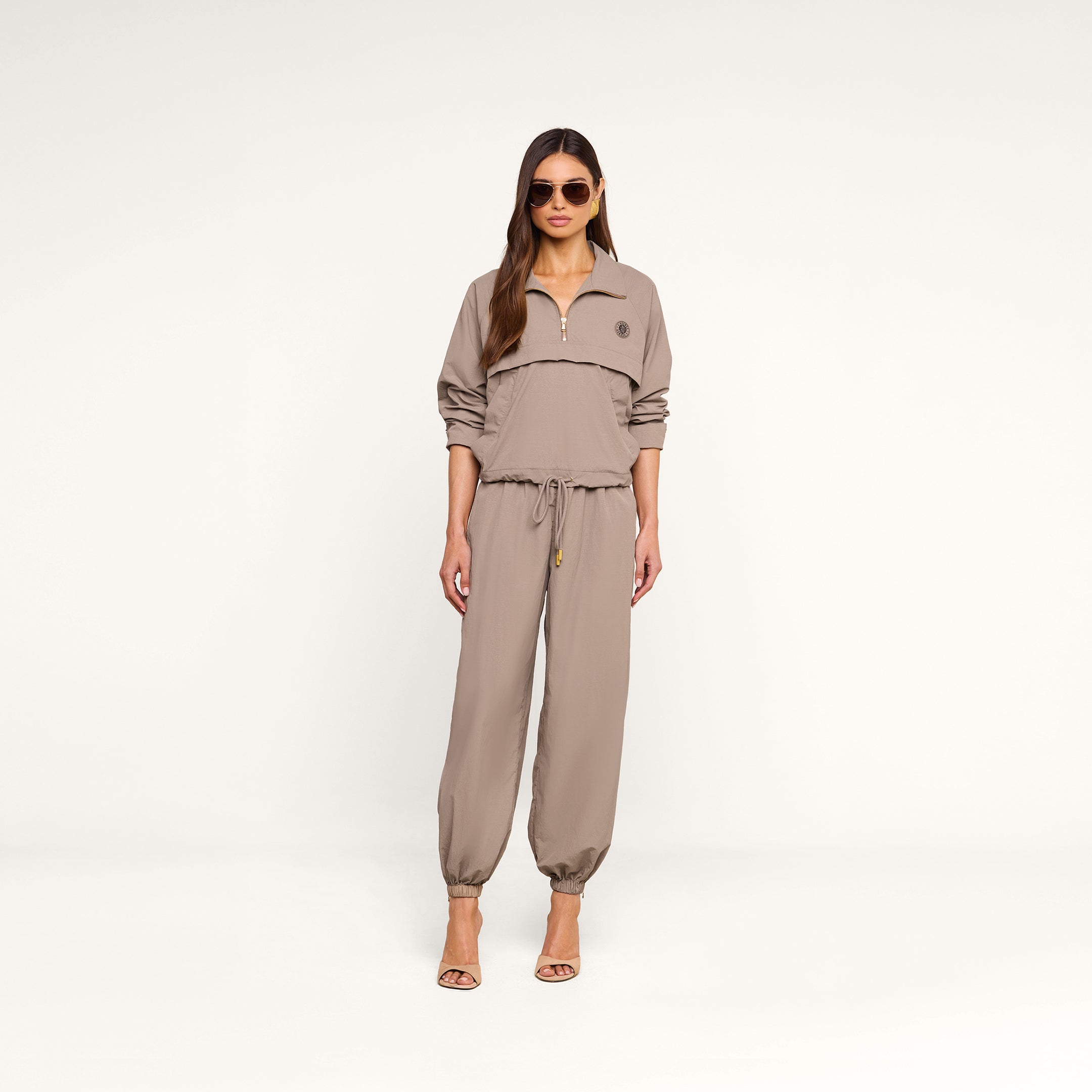 A woman stands against a plain background wearing sunglasses, the Vex Cropped Windbreaker in light brown, matching jogger pants, and heeled sandals.