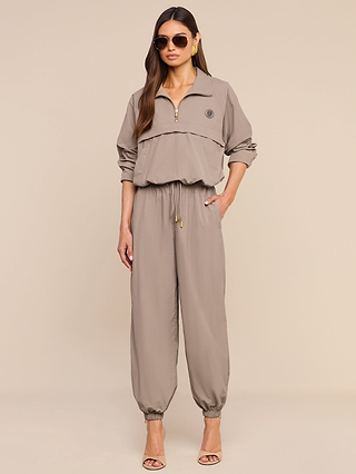 A woman wearing sunglasses and the Vex Cropped Windbreaker—a tan zip-up, long-sleeve jacket with elastic cuffs—stands with one hand in her pocket against a beige background, her look effortlessly elevated by the subtle water-repellent finish of this activewear-inspired style.