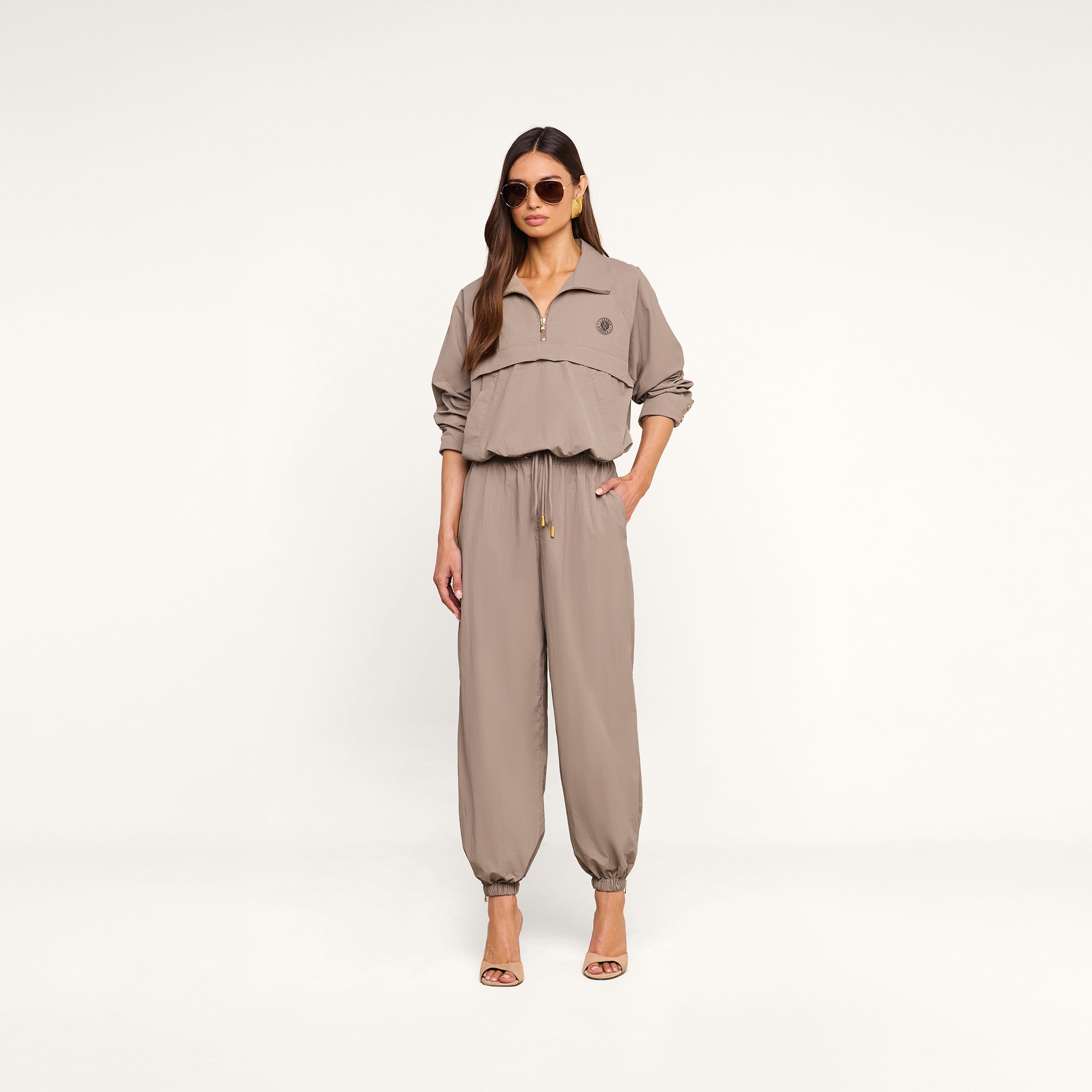A woman wearing sunglasses and the Vex Cropped Windbreaker with matching jogger pants and heeled sandals stands against a plain white background.