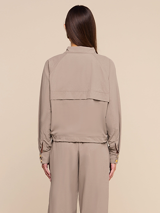 A woman with long brown hair, seen from behind, wears the Vex Cropped Windbreaker in beige along with matching pants against a plain light beige background.
