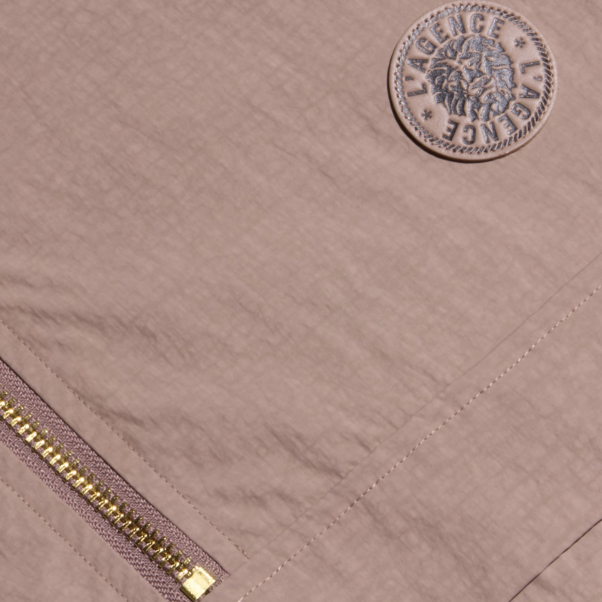 Close-up of the Vex Cropped Windbreaker, a mauve activewear jacket featuring a gold zipper and a round embroidered patch with a lion’s head and the word LAGENCE repeated twice around the border.