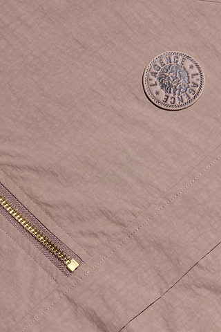 Close-up of the Vex Cropped Windbreaker in light mauve, showcasing its gold zipper and round patch with an emblem and multiple embossed “LAGENCE” details.