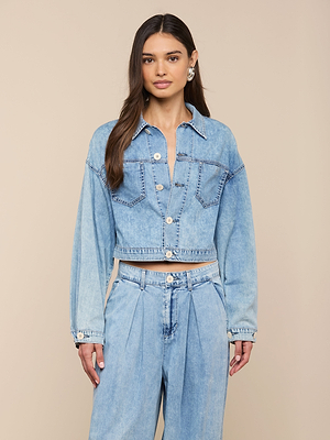 A woman with long, straight hair faces forward against a beige background, wearing the Gaines Jean Jacket in light blue featherweight denim with large buttons and matching high-waisted, pleated denim pants.