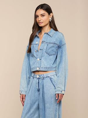 A woman with long brown hair wears the Gaines Jean Jacket, a featherweight denim button-up crop style, paired with matching high-waisted wide-leg jeans. She stands against a beige background, accessorized with silver hoop earrings.