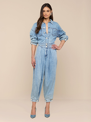 A woman with long dark hair and a neutral expression stands against a beige background wearing the Gaines Jean Jacket with rolled-up sleeves, belted waist, and blue high heels.