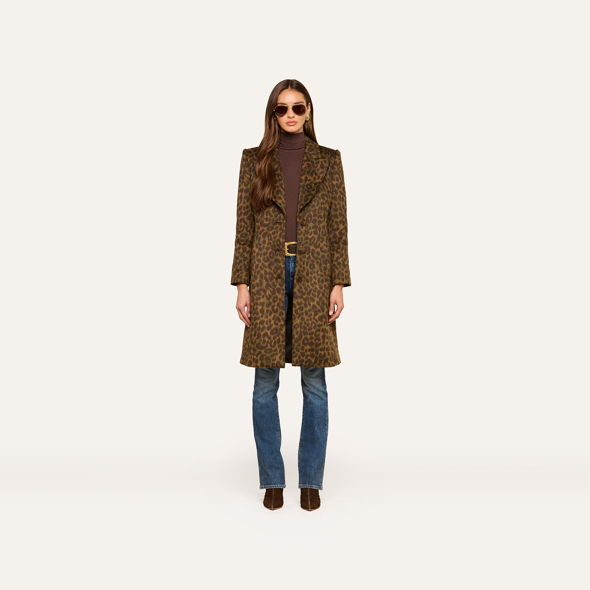 A woman wearing sunglasses, a brown turtleneck, blue jeans, brown ankle boots, and the Laney Coat stands against a plain white background.