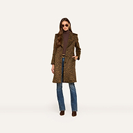 A woman stands against a plain light background wearing the Laney Coat, a long collared leopard print piece, paired with a brown turtleneck, blue jeans, and brown boots. She has long brown hair and sunglasses.