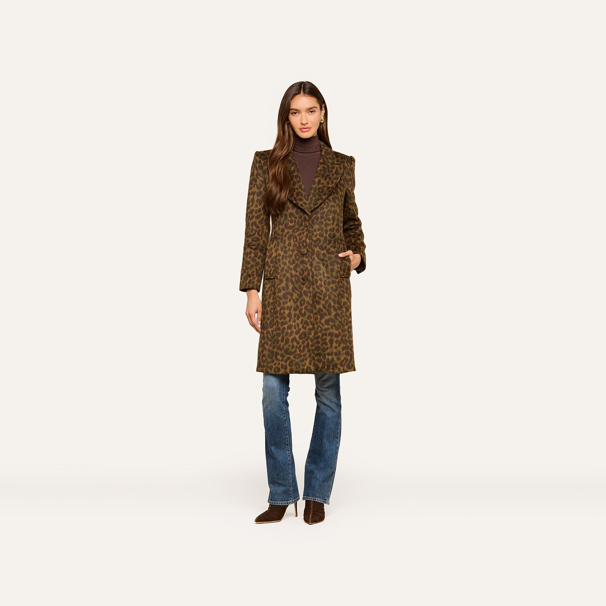 A woman stands facing forward with one hand in her pocket against a plain light background, wearing the Laney Coat—a long collared leopard print coat—paired with a brown turtleneck, blue jeans, and brown shoes.
