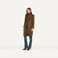 A woman stands against a plain, light background wearing the Laney Coat—a long, collared leopard print style—paired with a brown turtleneck, blue jeans, and brown shoes.