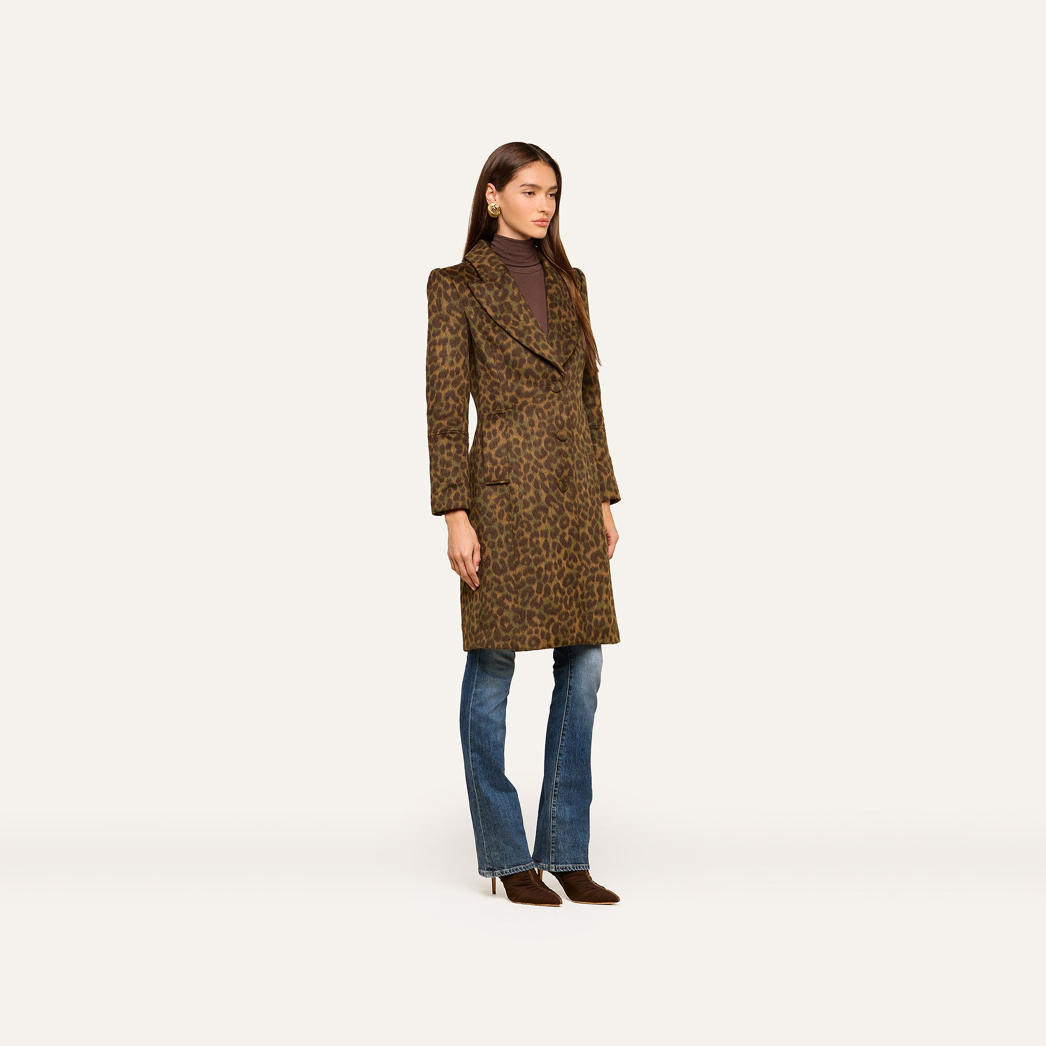 A woman stands against a plain white background wearing the Laney Coat—a long, collared leopard print design—layered over a brown turtleneck, blue jeans, and brown heels.
