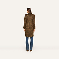 A woman stands facing away, wearing the Laney Coat—a long collared leopard print coat—paired with blue jeans and heeled shoes against a plain white background.