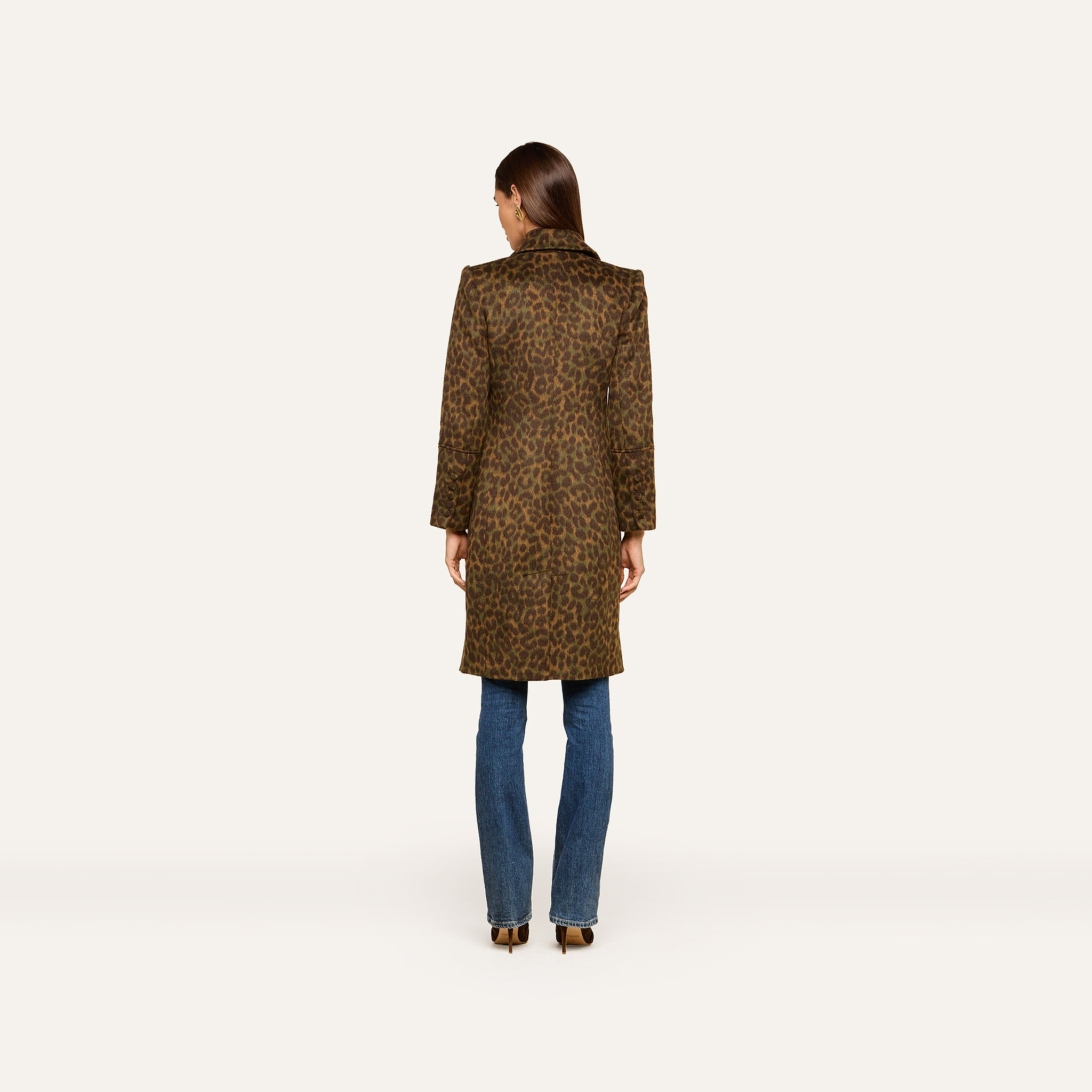 A woman stands facing away, wearing the Laney Coat—a long collared leopard print coat—paired with blue jeans and heeled shoes against a plain white background.