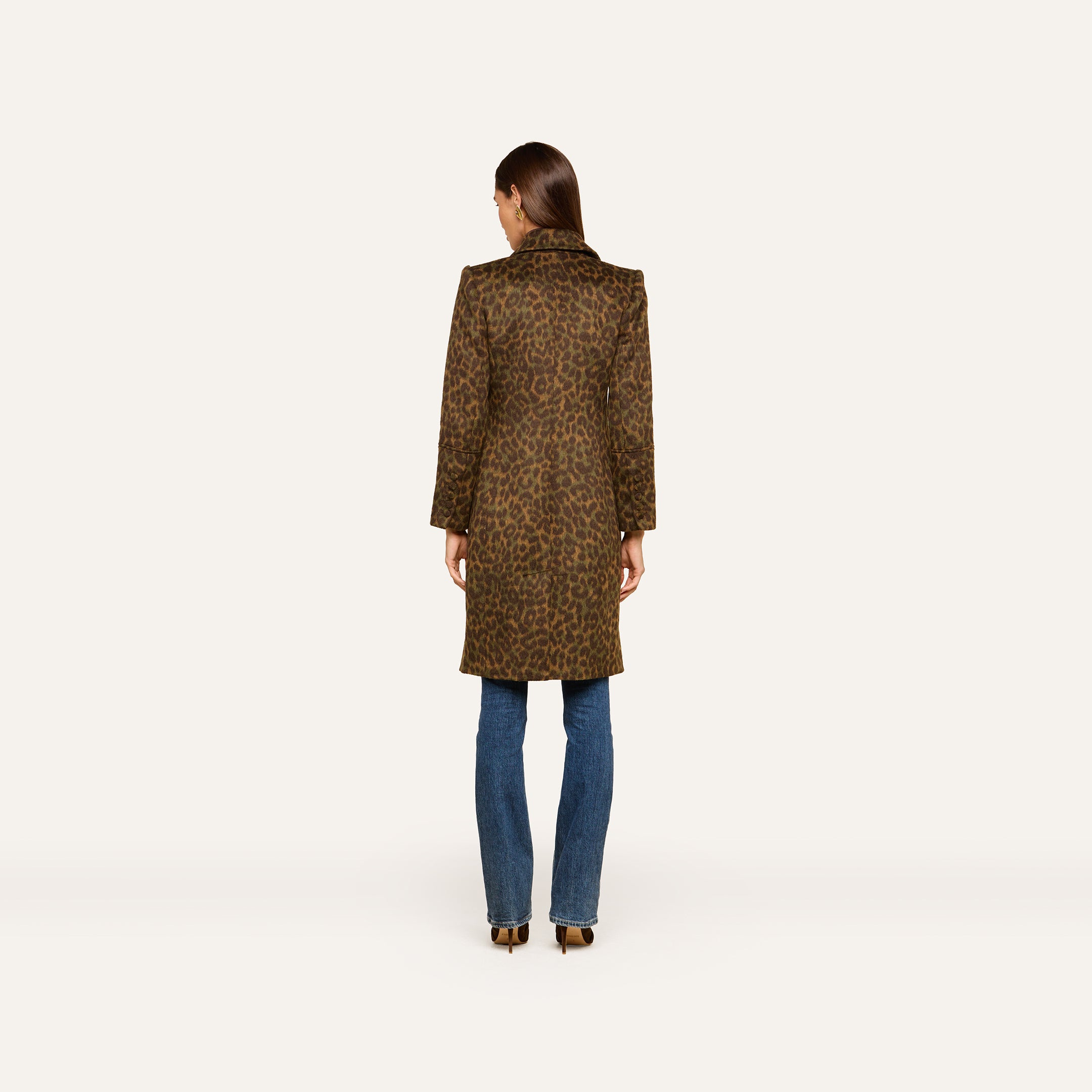 A woman stands facing away, wearing the Laney Coat—a long collared leopard print coat—paired with blue jeans and heeled shoes against a plain white background.