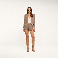 A woman wearing sunglasses, the Kahlia Jacket—a collarless lurex tweed piece with gold buttons—matching shorts, a light top, and knee-high tan boots stands against a plain white background, embodying quiet luxury.