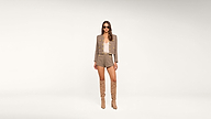 Woman stands facing forward wearing a taupe blazer with gold buttons, matching shorts, a light top, tall tan boots, and sunglasses against a plain white background.