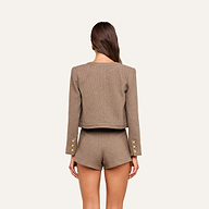 A woman stands with her back to the camera, wearing the collarless Kahlia Jacket in taupe, adorned with gold buttons and paired with matching high-waisted shorts. Her brown hair falls loose and straight against a plain off-white background, emphasizing the ensemble’s quiet luxury.