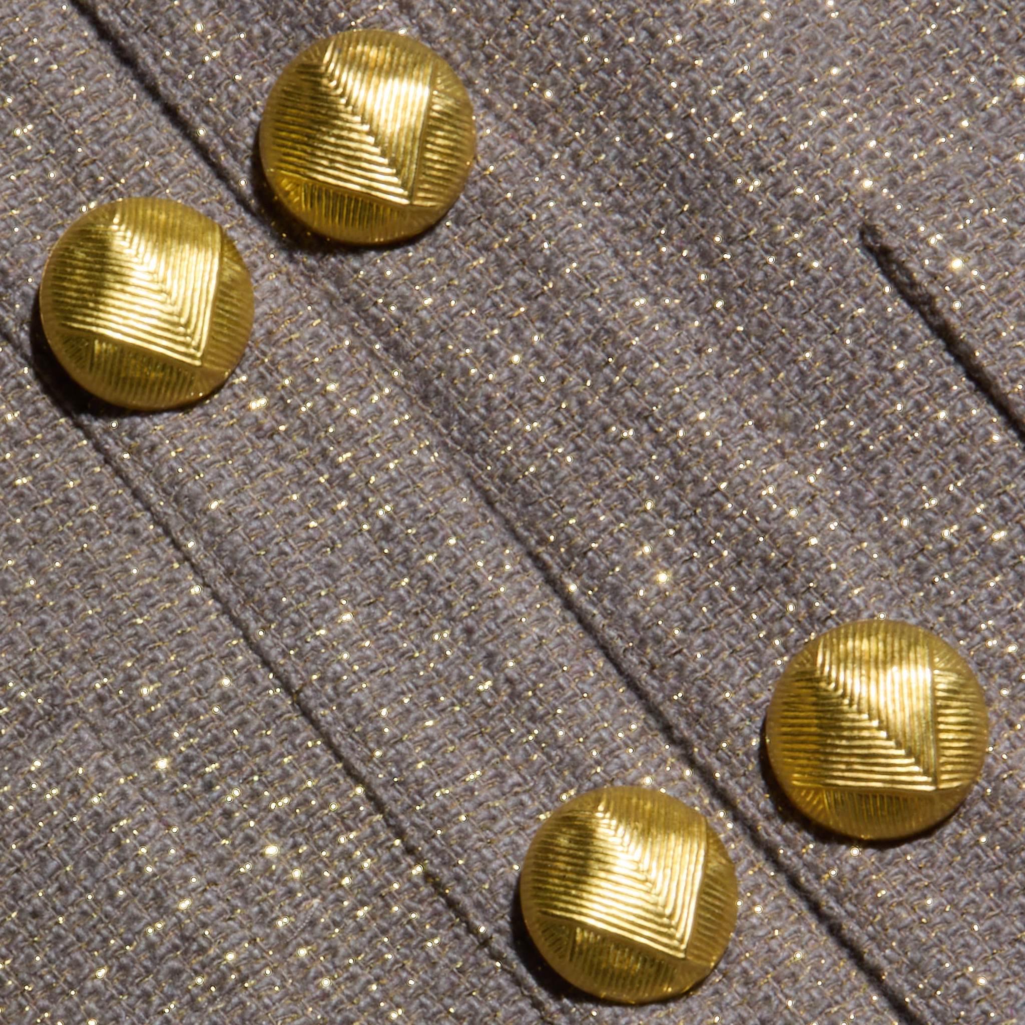 Four shiny gold buttons with a textured, geometric pattern are arranged on light brown sparkly lurex tweed fabric, featuring seam lines running diagonally across the surface—a detail that exemplifies quiet luxury in the Kahlia Jacket.