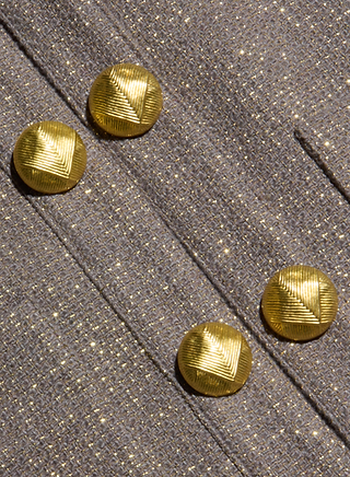 Four textured gold buttons are arranged diagonally on the shimmery gray lurex tweed fabric of the Kahlia Jacket, a collarless piece that embodies quiet luxury.