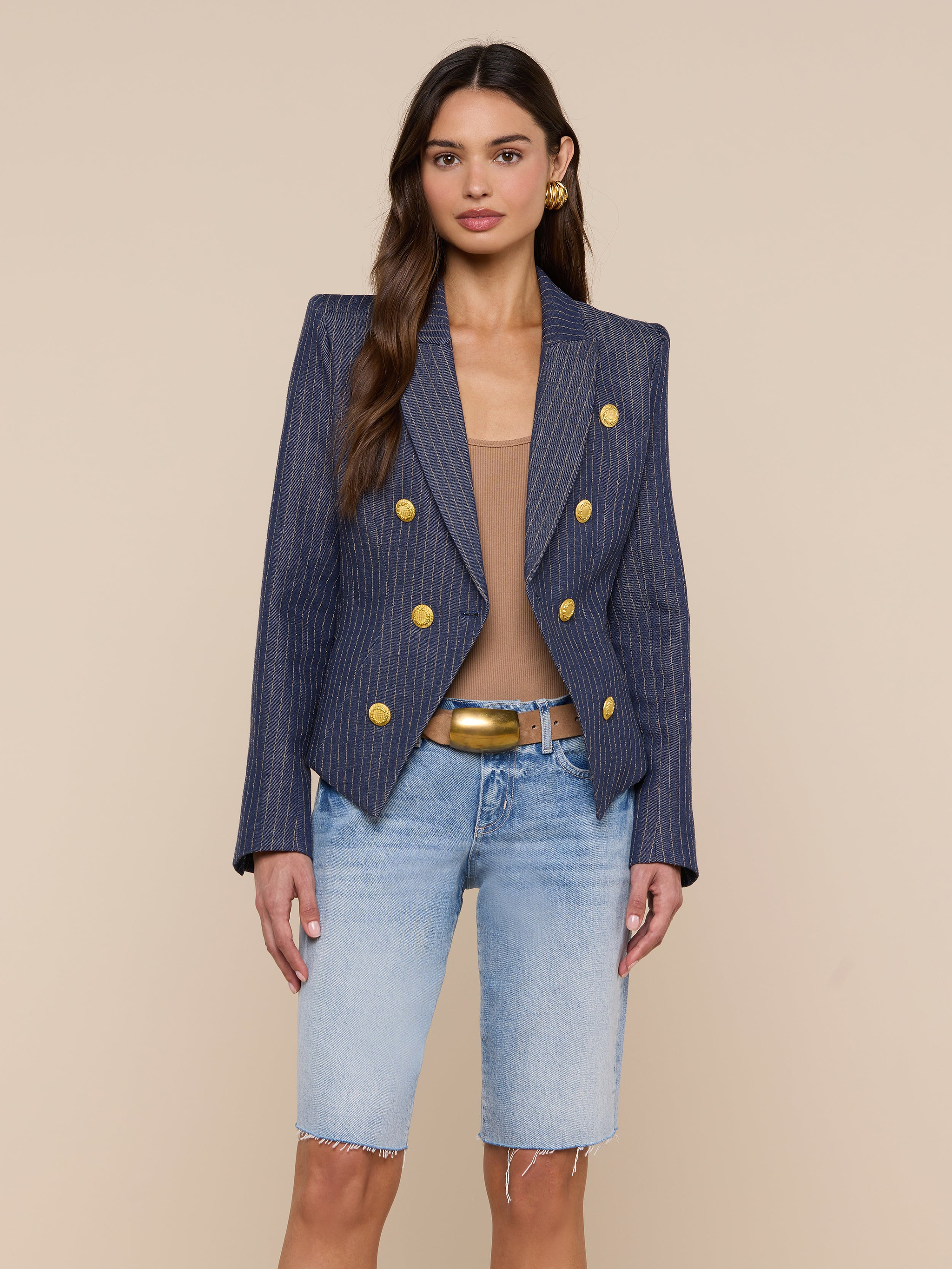 A woman wears the Solar Pinstriped Blazer—a slim fit, linen piece with gold buttons—paired with a tan top, denim Bermuda shorts, and a tan belt. She stands against a plain light background, looking at the camera.